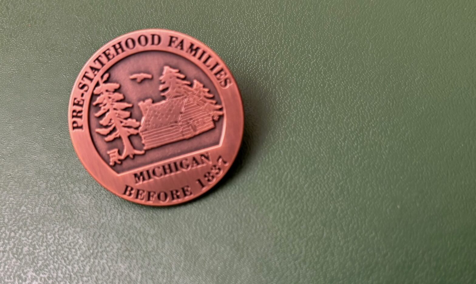 Circular copper-colored pin on a green background. Pin has the words “Pre-Statehood Families Michigan Before 1837” around the edge with symbols of a log cabin and white pines in the center.
